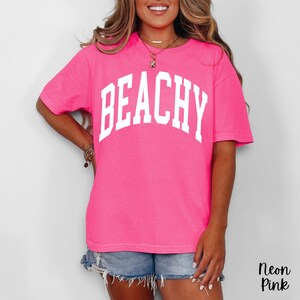 May include: A neon pink t-shirt with the word "BEACHY" printed in large, white, block letters across the front. The shirt is a vibrant, solid color and has short sleeves. The text "Neon Pink" is in the bottom right corner.