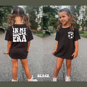 May include: Black t-shirt with white text that says "In My Soccer Era" on the back and a white soccer ball with the name "Olivia" on the front.