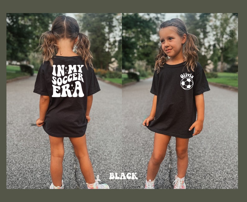 May include: Black t-shirt with white text that says "In My Soccer Era" on the back and a white soccer ball with the name "Olivia" on the front.