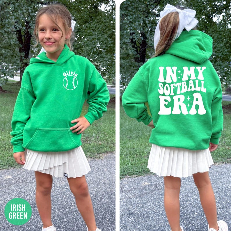 May include: A green hooded sweatshirt with a white softball graphic and the name "Olivia". The back of the sweatshirt says "IN MY SOFTBALL ERA" in white. The person is wearing a white pleated skirt. The words "Irish Green" are at the bottom.
