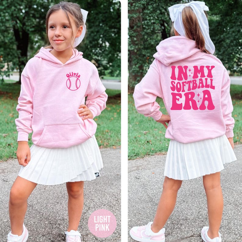 May include: A light pink hoodie with a front pocket and a white pleated skirt. The hoodie has a baseball graphic with the name "Olivia" and the text "IN MY SOFTBALL ERA" on the back. The outfit is paired with white sneakers.