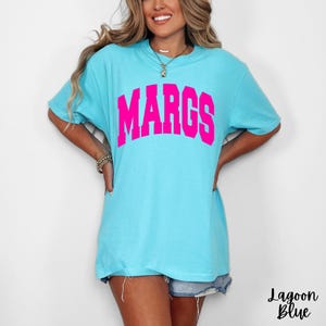 May include: A light blue t-shirt with the word "MARGS" in bright pink block letters across the chest. The shirt is a relaxed fit and has short sleeves. The words "Lagoon Blue" are written in cursive at the bottom right.