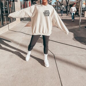 Puede incluir: Una mujer lleva una sudadera de color crema con el texto "Anti Social Wives Club" en letras negras. Lleva leggings negros y zapatillas blancas.