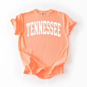 K&ouml;nnte beinhalten: Ein pfirsichfarbenes T-Shirt mit dem Wort "TENNESSEE" in wei&szlig; auf der Vorderseite.