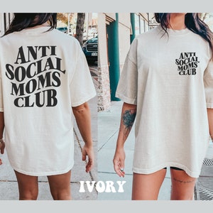 May include: A white oversized t-shirt with black text that reads "Anti Social Moms Club". The shirt is shown on a model in an outdoor setting. The model is wearing the shirt with the text on the back.