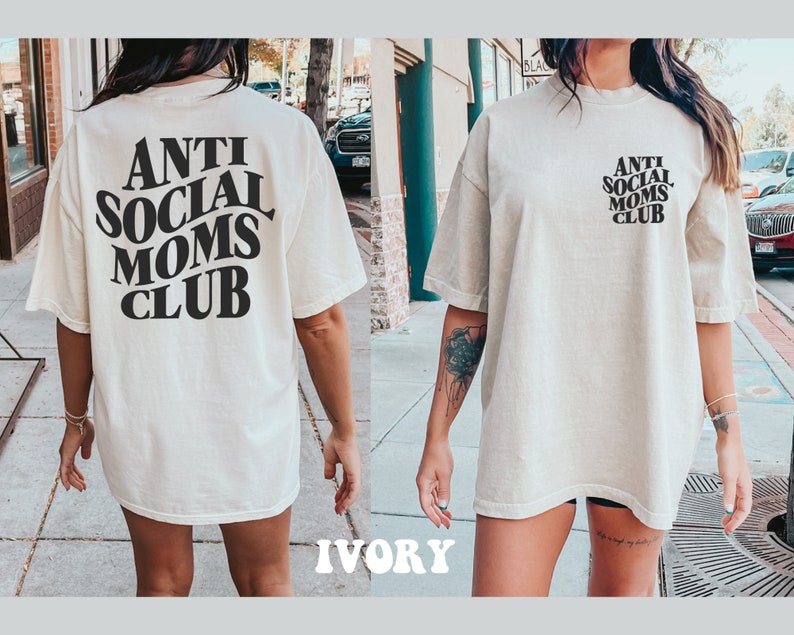 May include: A white oversized t-shirt with black text that reads "Anti Social Moms Club". The shirt is shown on a model in an outdoor setting. The model is wearing the shirt with the text on the back.