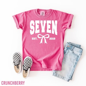 May include: A pink t-shirt with the word "SEVEN" in white block letters, a white bow, and the text "EST. 2019". The shirt is paired with light blue denim shorts and black and white checkered slip-on shoes.