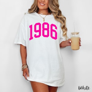 May include: White t-shirt with the year "1986" printed in bright pink block letters. The shirt is a casual, oversized style. The word "White" is printed in the bottom right corner.