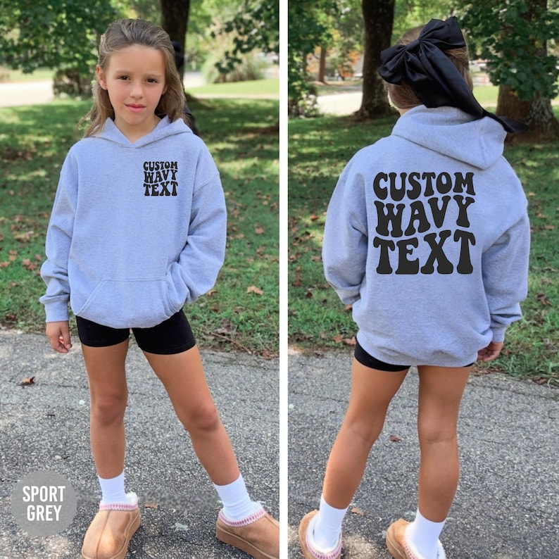 Puede incluir: Una sudadera con capucha gris claro con el texto "CUSTOM WAVY TEXT" impreso en negro en la parte delantera y trasera. La sudadera tiene un bolsillo delantero y una capucha. La esquina inferior izquierda de la imagen tiene el texto "SPORT GREY".