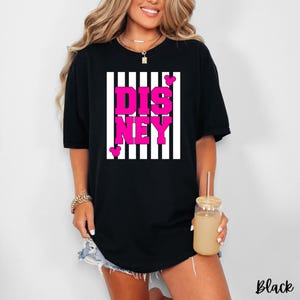 Może przedstawiać: Czarna koszulka z napisem "DISNEY" w jaskrawor&oacute;żowych literach, na tle białych pask&oacute;w. Koszulka ma luźny kr&oacute;j i kr&oacute;tkie rękawy. Małe ikony głowy Myszki Miki są obecne.