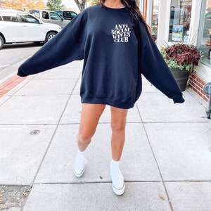 Puede incluir: Una sudadera negra con el texto "ANTI SOCIAL WIVES CLUB" en letras blancas. La sudadera la lleva puesta una persona con zapatillas blancas.