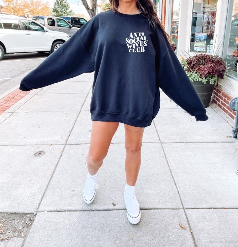 Puede incluir: Una sudadera negra con el texto "ANTI SOCIAL WIVES CLUB" en letras blancas. La sudadera la lleva puesta una persona con zapatillas blancas.
