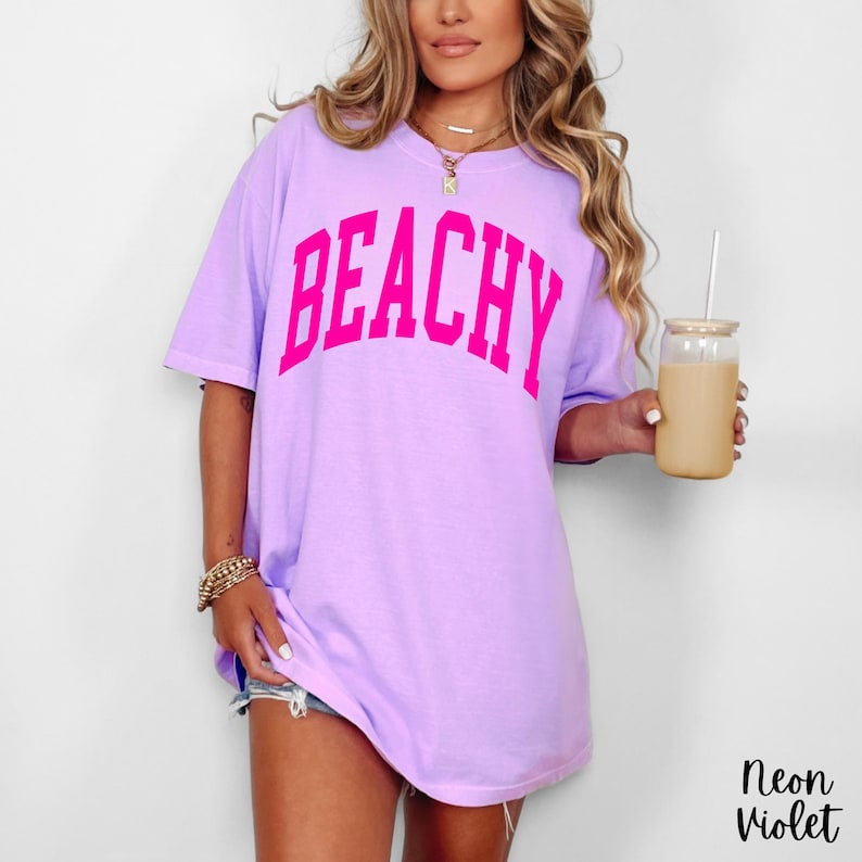 May include: A light purple t-shirt with the word "BEACHY" in bright pink block letters. The shirt is oversized and has short sleeves. The model is holding a glass of iced coffee. The text "Neon Violet" is in the bottom right corner.