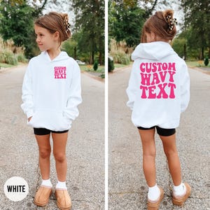 Puede incluir: Una sudadera con capucha blanca con el texto rosa "CUSTOM WAVY TEXT" en la parte delantera y trasera. La sudadera tiene un bolsillo delantero y una capucha. La modelo lleva pantalones cortos negros y zapatos color canela. La palabra "WHITE" est&aacute; en un c&iacute;rculo blanco en la parte inferior.