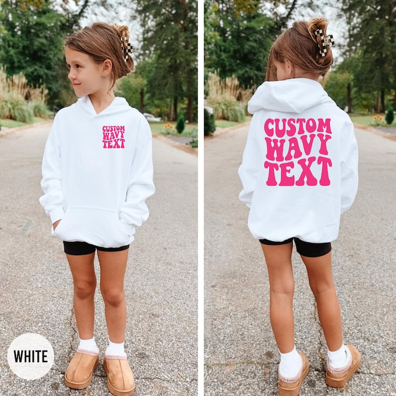 Puede incluir: Una sudadera con capucha blanca con el texto rosa "CUSTOM WAVY TEXT" en la parte delantera y trasera. La sudadera tiene un bolsillo delantero y una capucha. La modelo lleva pantalones cortos negros y zapatos color canela. La palabra "WHITE" est&aacute; en un c&iacute;rculo blanco en la parte inferior.
