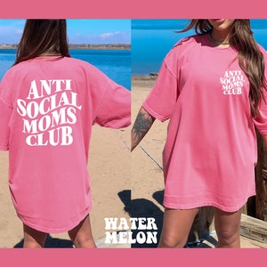 May include: Pink oversized t-shirt with white text that reads "Anti Social Moms Club". The shirt is shown on a model standing on a beach.