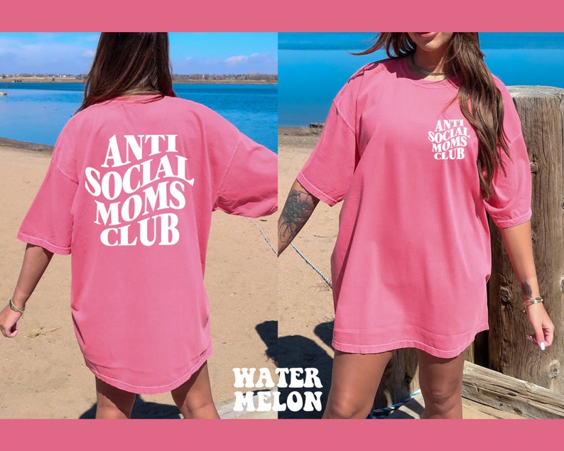 May include: Pink oversized t-shirt with white text that reads "Anti Social Moms Club". The shirt is shown on a model standing on a beach.