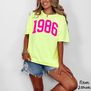 May include: A neon yellow t-shirt with the year "1986" printed in bright pink block letters. The shirt is paired with denim shorts. The words "Neon Lemon" are printed in black at the bottom right corner.
