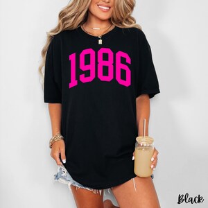 May include: Black t-shirt with the number "1986" in bright pink block letters. The word "Black" is printed in the bottom right corner. The shirt has short sleeves and a round neckline.
