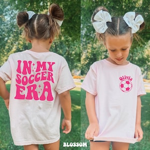 May include: Pink t-shirt with a pink graphic that says "In My Soccer Era" on the back and a pink soccer ball with the name "Olivia" on the front.
