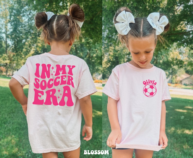 May include: Pink t-shirt with a pink graphic that says "In My Soccer Era" on the back and a pink soccer ball with the name "Olivia" on the front.
