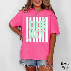 Może przedstawiać: Neonowo-r&oacute;żowa koszulka z biało-miętowym nadrukiem Disneya. Grafika przedstawia napis "DISNEY" w miętowych literach blokowych na białym, pasiastym tle. Na grafice znajdują się uszy Myszki Miki. Koszulka jest koszulką z kr&oacute;tkim rękawem.