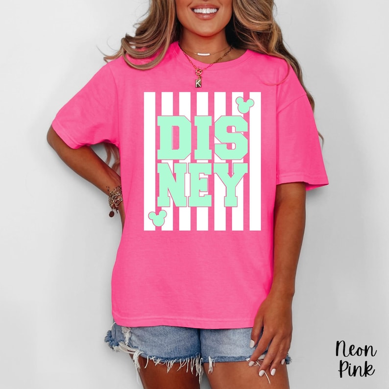 Może przedstawiać: Neonowo-r&oacute;żowa koszulka z biało-miętowym nadrukiem Disneya. Grafika przedstawia napis "DISNEY" w miętowych literach blokowych na białym, pasiastym tle. Na grafice znajdują się uszy Myszki Miki. Koszulka jest koszulką z kr&oacute;tkim rękawem.