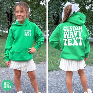 Puede incluir: Una sudadera con capucha verde con el texto "CUSTOM WAVY TEXT" en blanco. La sudadera tiene un bolsillo delantero y se combina con una falda plisada blanca. Las palabras "IRISH GREEN" est&aacute;n en la parte inferior de la imagen.