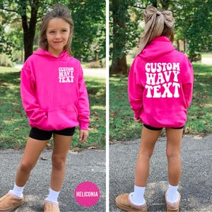 Puede incluir: Una sudadera con capucha rosa brillante con el texto blanco "CUSTOM WAVY TEXT" en la parte delantera y trasera. La sudadera se combina con pantalones cortos negros, calcetines blancos y zapatos marrones. El fondo es un parque.