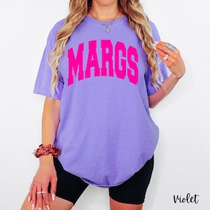 May include: A violet-colored t-shirt with the word "MARGS" printed in bright pink across the chest. The shirt has short sleeves and a relaxed fit. The word "Violet" is printed in a script font at the bottom right corner.
