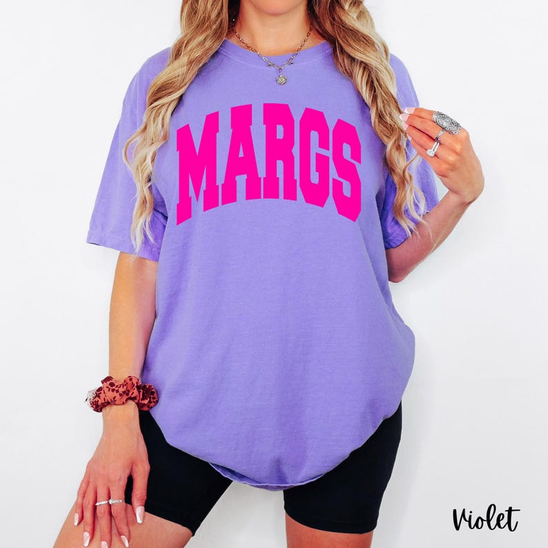 May include: A violet-colored t-shirt with the word "MARGS" printed in bright pink across the chest. The shirt has short sleeves and a relaxed fit. The word "Violet" is printed in a script font at the bottom right corner.