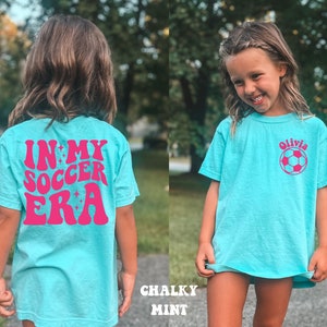 May include: A turquoise blue t-shirt with a pink graphic that says "In My Soccer Era" on the back. The front of the shirt has a pink soccer ball and the name "Olivia" printed on it.