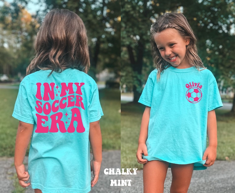 May include: A turquoise blue t-shirt with a pink graphic that says "In My Soccer Era" on the back. The front of the shirt has a pink soccer ball and the name "Olivia" printed on it.