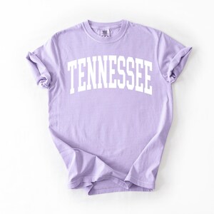K&ouml;nnte beinhalten: Ein lavendelfarbenes T-Shirt mit dem Wort "TENNESSEE" in wei&szlig; in einem Bogen &uuml;ber der Vorderseite.