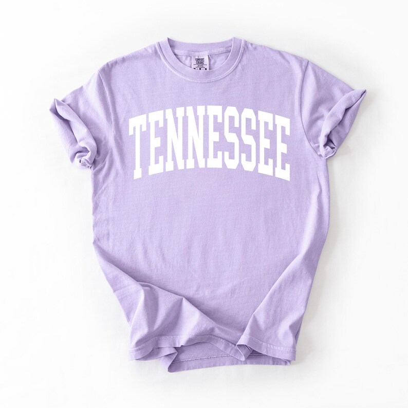 K&ouml;nnte beinhalten: Ein lavendelfarbenes T-Shirt mit dem Wort "TENNESSEE" in wei&szlig; in einem Bogen &uuml;ber der Vorderseite.