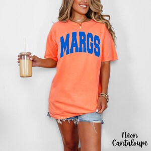 May include: A neon cantaloupe-colored t-shirt with the word "MARGS" in blue block letters. The shirt is paired with denim shorts. A glass tumbler with a straw is held in the left hand. The text "Neon Cantaloupe" is in the bottom right corner.