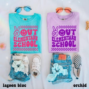 May include: Two t-shirts, one lagoon blue and one orchid, with the text "OUT ELEMENTARY SCHOOL" in a retro font. The shirts are paired with denim shorts, scrunchies, sunglasses, and trainers. The lagoon blue shirt has pink text, and the orchid shirt has purple text.