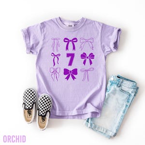May include: A light purple t-shirt with a design of nine purple bows and the number 7. The shirt is paired with checkered black and white slip-on shoes and light blue denim shorts. The word "ORCHID" is at the bottom.