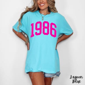 May include: Lagoon blue t-shirt with the year "1986" printed in bright pink. The shirt has short sleeves and a relaxed fit. The text "Lagoon Blue" is printed in cursive at the bottom right.