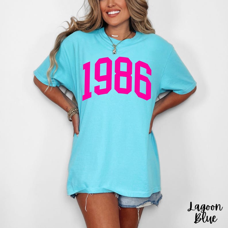 May include: Lagoon blue t-shirt with the year "1986" printed in bright pink. The shirt has short sleeves and a relaxed fit. The text "Lagoon Blue" is printed in cursive at the bottom right.