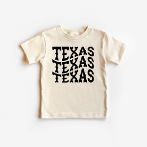 K&ouml;nnte beinhalten: Ein cremefarbenes T-Shirt mit kurzen &Auml;rmeln, auf dem das Wort "TEXAS" viermal in einer schwarzen, stilisierten Schrift wiederholt wird.