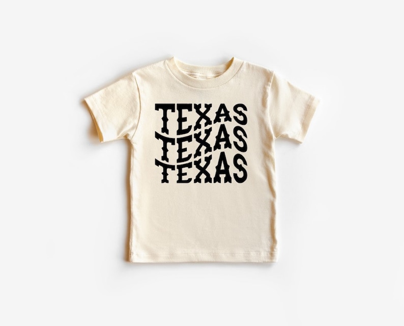 K&ouml;nnte beinhalten: Ein cremefarbenes T-Shirt mit kurzen &Auml;rmeln, auf dem das Wort "TEXAS" viermal in einer schwarzen, stilisierten Schrift wiederholt wird.