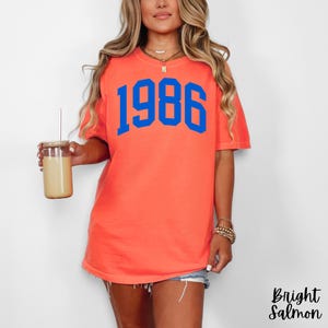 May include: A coral-colored t-shirt with the blue number "1986" printed on the front. The shirt is a short-sleeved, casual style. The words "Bright Salmon" are printed in a script font on the bottom right corner of the image.