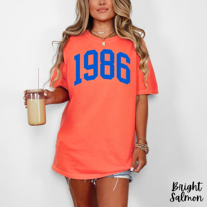 May include: A coral-colored t-shirt with the blue number "1986" printed on the front. The shirt is a short-sleeved, casual style. The words "Bright Salmon" are printed in a script font on the bottom right corner of the image.