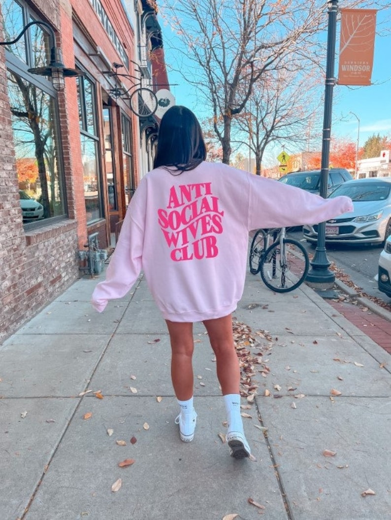 Puede incluir: Una sudadera rosa con el texto "Anti Social Wives Club" en letras rosas en la parte posterior.