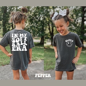 May include: Two children wearing dark grey t-shirts with white text. The back of the shirt says "In My Soft Ball Era" with stars. The front of the shirt says "Olivia" with a smiley face and hearts.