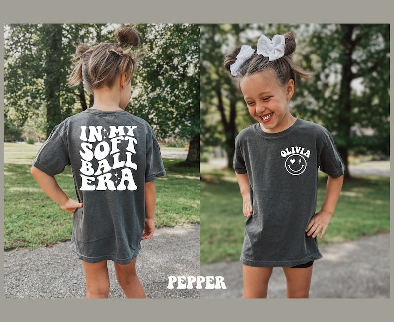 May include: Two children wearing dark grey t-shirts with white text. The back of the shirt says "In My Soft Ball Era" with stars. The front of the shirt says "Olivia" with a smiley face and hearts.