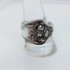 May include: A silver ring with a floral design. The ring features a detailed flower in the center, with intricate patterns on the band. The ring is displayed on a clear stand, showcasing its craftsmanship and design.