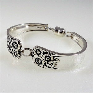 May include: Silver spoon bracelet with floral design and a magnetic clasp. The bracelet is marked "Wallace" and "Grand" on the back.