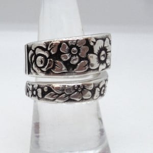 May include: A silver-colored ring with a floral design. The ring features detailed carvings of flowers and leaves, wrapping around the finger. The ring is displayed on a clear, conical stand.